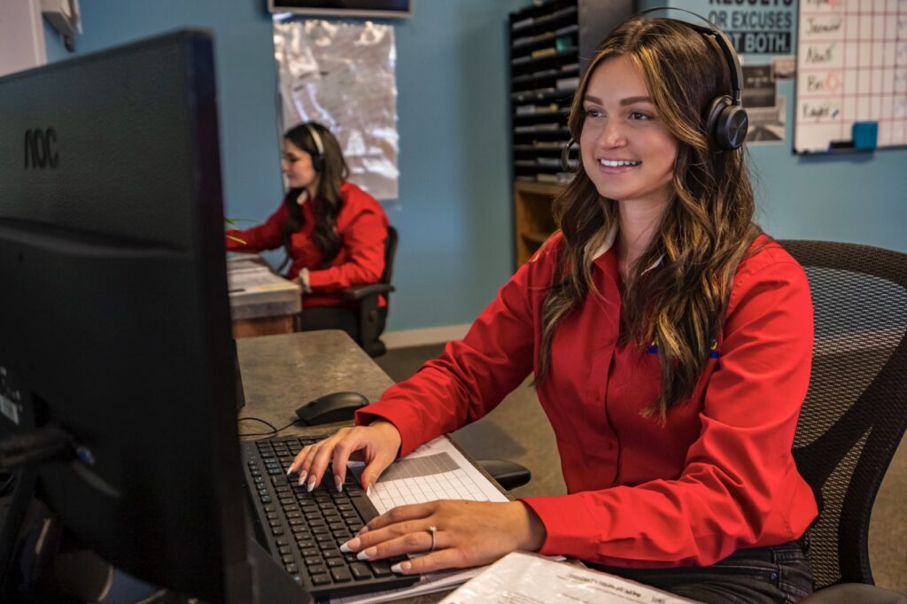 Advanced Home Services appointment scheduler sitting at a desk, wearing red button-up shirt and a headset while typing at a desktop computer.